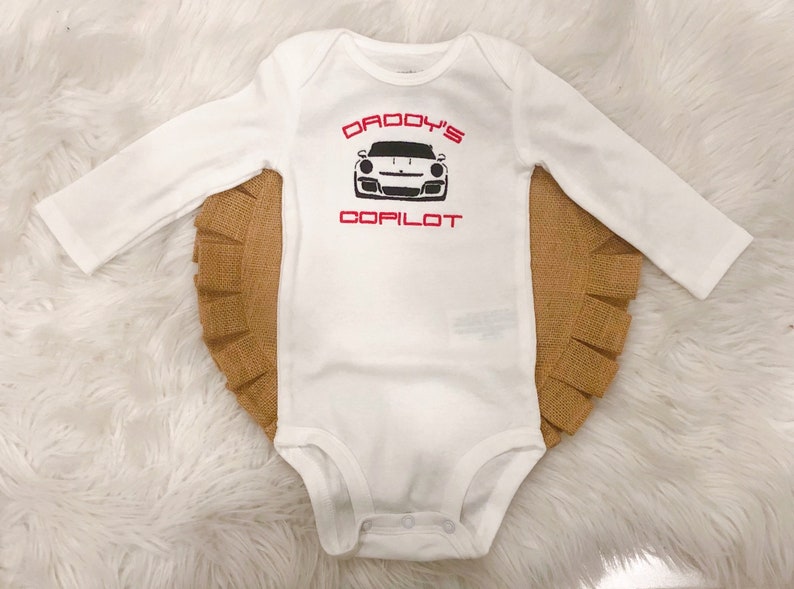 May include: White long-sleeved baby onesie with red text that says "Daddy's Copilot" and a black and white image of a sports car.