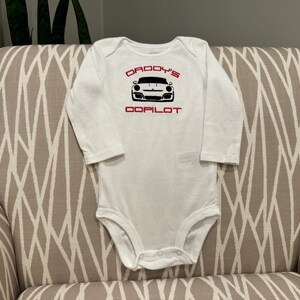 May include: White long-sleeve baby onesie with red text that reads "Daddy's Copilot" and a black and white image of a sports car.