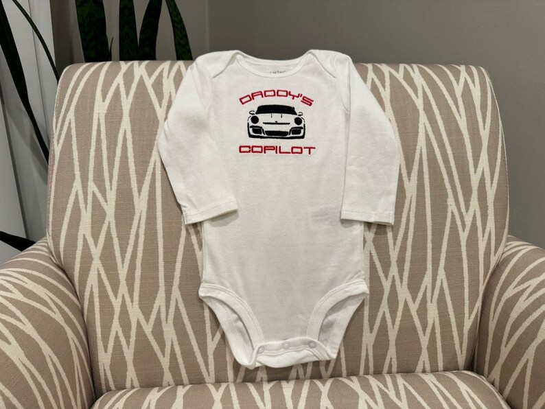 May include: White long-sleeve baby onesie with red text that reads "Daddy's Copilot" and a black and white image of a sports car.