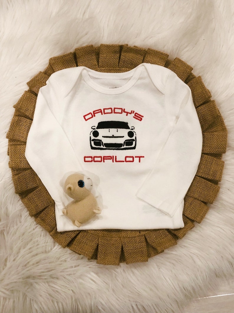 Puede incluir: Mono blanco de manga larga para beb&eacute; con texto rojo que dice "Daddy's Copilot" y una imagen en blanco y negro de un coche deportivo.