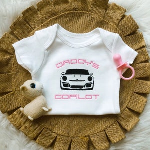 Puede incluir: Un body blanco para beb&eacute; con la inscripci&oacute;n "Daddy's Copilot" en rosa y una imagen en blanco y negro de un coche deportivo.