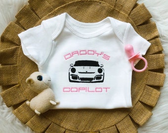 May include: White baby onesie with pink lettering that says "Daddy's Copilot" and a black and white image of a sports car.