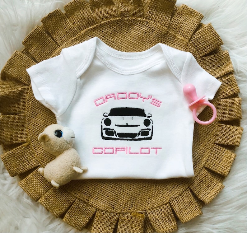 Puede incluir: Un body blanco para beb&eacute; con la inscripci&oacute;n "Daddy's Copilot" en rosa y una imagen en blanco y negro de un coche deportivo.