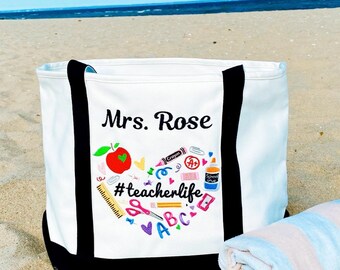 May include: White canvas tote bag with black handles and a black trim. The bag has an embroidered design of a red apple, school supplies, and the text "#teacherlife". The name "Mrs. Rose" is embroidered above the design.