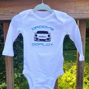 Puede incluir: Mono blanco de manga larga para beb&eacute; con bordado azul y negro. El bordado dice "Daddy's Copilot" y presenta una silueta negra de un coche deportivo.