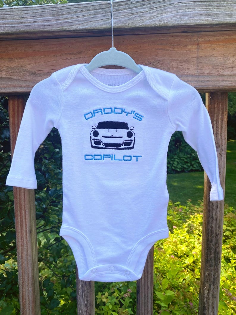 Puede incluir: Mono blanco de manga larga para beb&eacute; con bordado azul y negro. El bordado dice "Daddy's Copilot" y presenta una silueta negra de un coche deportivo.