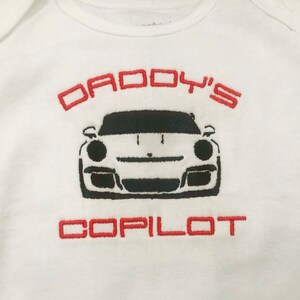 Puede incluir: Uniforme blanco para beb&eacute; con el texto bordado en rojo "Daddy's Copilot" y una silueta negra de un coche deportivo.