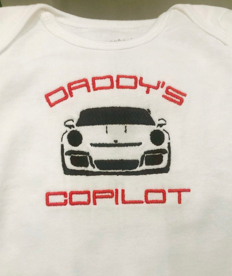 Puede incluir: Uniforme blanco para beb&eacute; con el texto bordado en rojo "Daddy's Copilot" y una silueta negra de un coche deportivo.