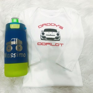 Puede incluir: Un mono blanco de manga larga con el texto "Daddy's Copilot" y una imagen en blanco y negro de un coche deportivo. Una botella de agua azul y verde con un gr&aacute;fico de un cami&oacute;n monstruo y el nombre "Massimo" est&aacute; en primer plano.