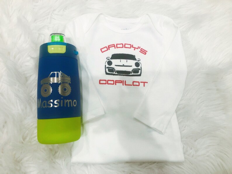 Puede incluir: Un mono blanco de manga larga con el texto "Daddy's Copilot" y una imagen en blanco y negro de un coche deportivo. Una botella de agua azul y verde con un gr&aacute;fico de un cami&oacute;n monstruo y el nombre "Massimo" est&aacute; en primer plano.