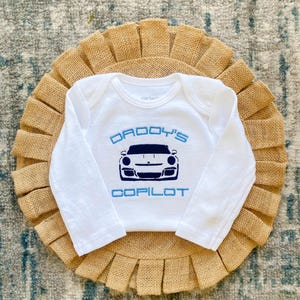 Puede incluir: Mono blanco de manga larga para beb&eacute; con bordado azul. El mono dice "Copiloto de pap&aacute;" y presenta una imagen de dibujos animados de un coche deportivo blanco.