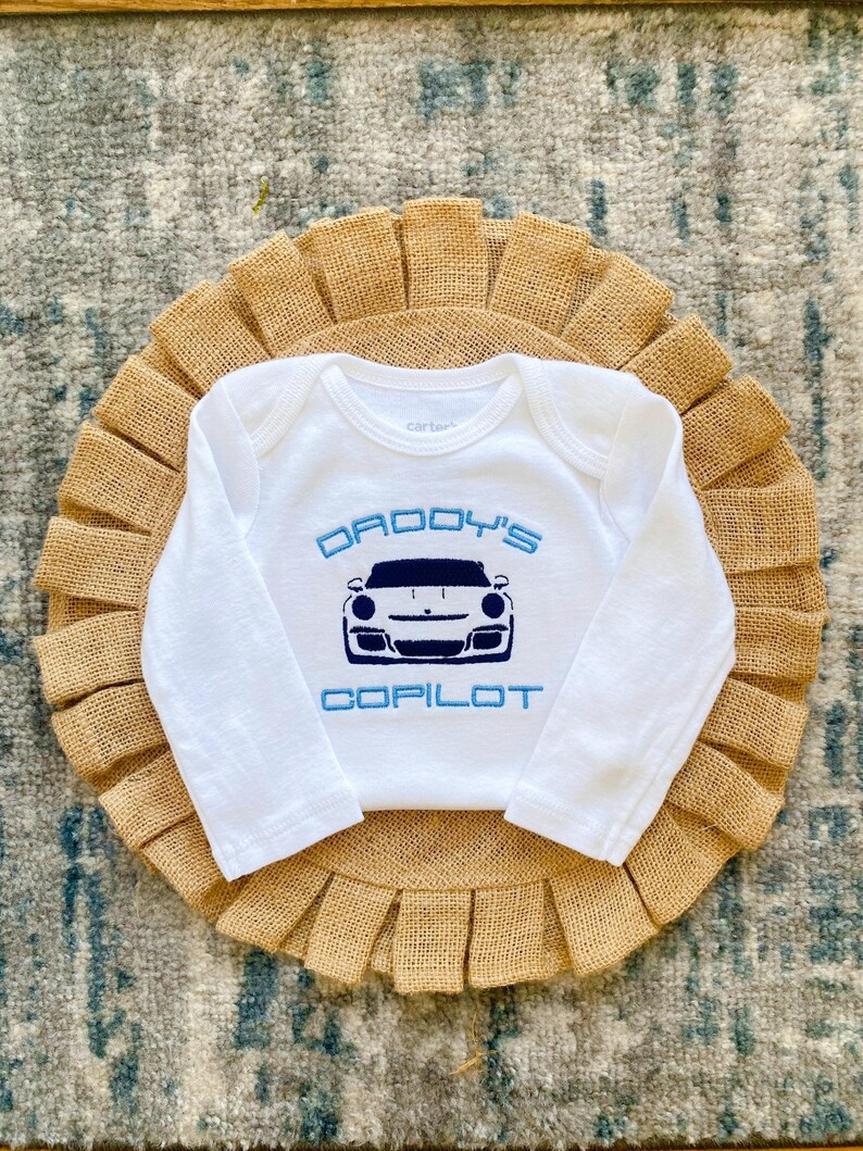 Puede incluir: Mono blanco de manga larga para beb&eacute; con bordado azul. El mono dice "Copiloto de pap&aacute;" y presenta una imagen de dibujos animados de un coche deportivo blanco.