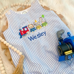 May include: A light blue and white striped sleeveless romper featuring an embroidered train with a lion, elephant, and giraffe wearing party hats. The train engine is red with the number one, and the name Wesley is embroidered in blue script.