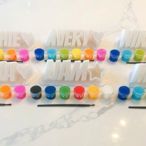 May include: A set of six paint-your-own name signs with paint pots and brushes. The names are Sophie, Emma, Avery, Liam, Nikki, and Mia.