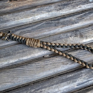 Könnte beinhalten: Ein schwarzes und braunes Paracord-Lanyard mit einem Metallverschluss. Das Lanyard liegt auf einer Holzoberfläche.