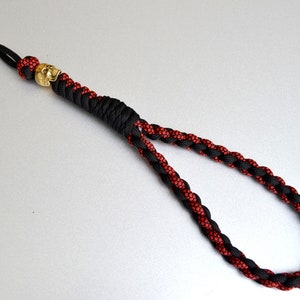 May include: A black and red braided lanyard with a gold skull bead. The lanyard has a loop at one end and a long, thin, braided cord.