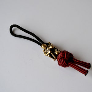 Può includere: Un cordoncino nero con un cordoncino intrecciato rosso e nero attaccato a un ciondolo in metallo dorato. Il ciondolo è un design dettagliato di due figure.