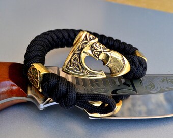 May include: A black paracord bracelet with a gold-toned Viking axe charm. The bracelet is lying on top of a brown and silver knife.