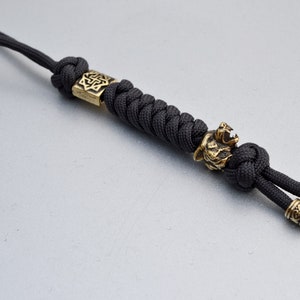 May include: Black paracord bracelet with gold metal beads. The beads feature a wolf head design and a Celtic knot design.