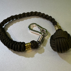May include: A black paracord keychain with a gold bead and a silver carabiner. The keychain has a black weighted ball at the end.