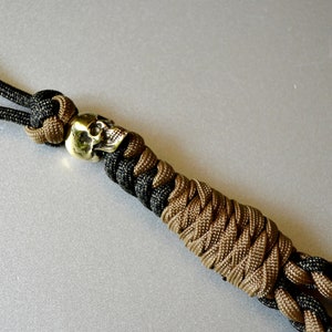 May include: A black and brown paracord bracelet with a brass skull bead. The bracelet is made with a cobra knot.