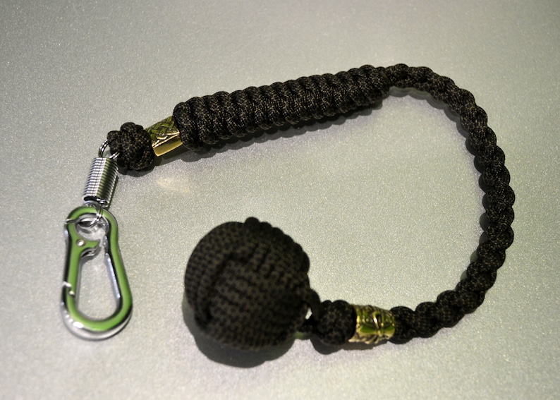 May include: A black paracord keychain with a metal carabiner clip and a black weighted ball at the end. The keychain has two gold metal beads.