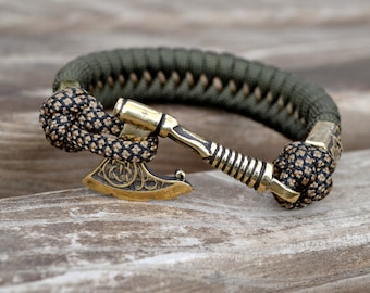 Op de afbeelding: Een groene paracord armband met een bronzen bijl bedel. De bijl bedel heeft ingewikkelde details en is met een zwart-bruine knoop aan de armband bevestigd. De armband is ontworpen om om de pols te worden gedragen.