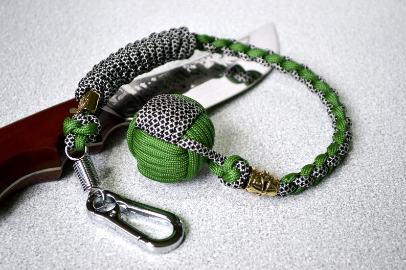 May include: A green and black paracord keychain with a metal carabiner, a spring, and a small green and black ball. The keychain is attached to a brown and silver knife.