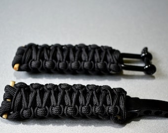 May include: Two black paracord keychains with metal attachments. The keychain on the left has a simple loop attachment, while the keychain on the right has a more complex attachment with multiple holes.
