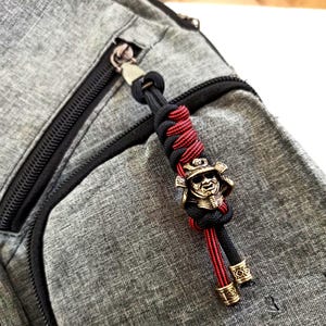 May include: A black and red braided lanyard with a bronze-colored samurai helmet charm. The lanyard is attached to a gray backpack with black zippers. The samurai charm has intricate details.
