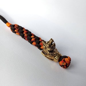 May include: A black and orange paracord bracelet with a wolf head charm. The charm is made of brass and has a detailed design.