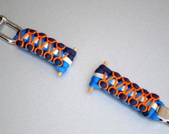 May include: A blue, white, and orange paracord bracelet with a silver metal clasp. The bracelet is made with a knotting technique and has a decorative pattern.