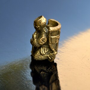 以下が含まれることがあります： 鎧を着た騎士の金色の金属製フィギュアで、剣を手にひざまずいています。騎士は兜をかぶり、背中に盾を背負っています。