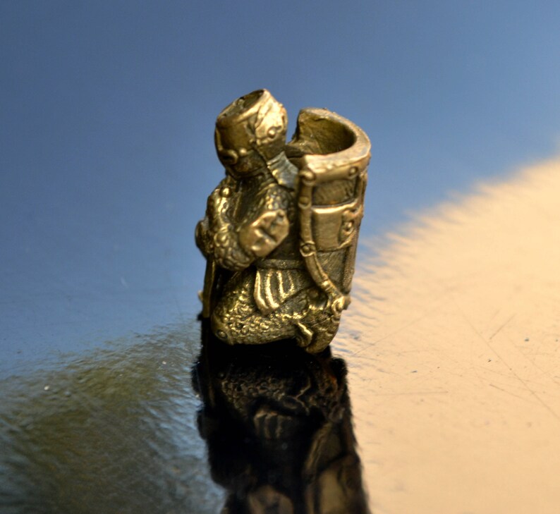 以下が含まれることがあります： 鎧を着た騎士の金色の金属製フィギュアで、剣を手にひざまずいています。騎士は兜をかぶり、背中に盾を背負っています。