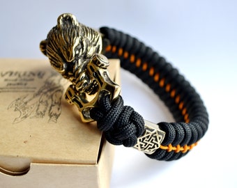 Puede incluir: Un brazalete de paracord negro y naranja con un dije de cabeza de oso de latón y un dije de nudo de latón.