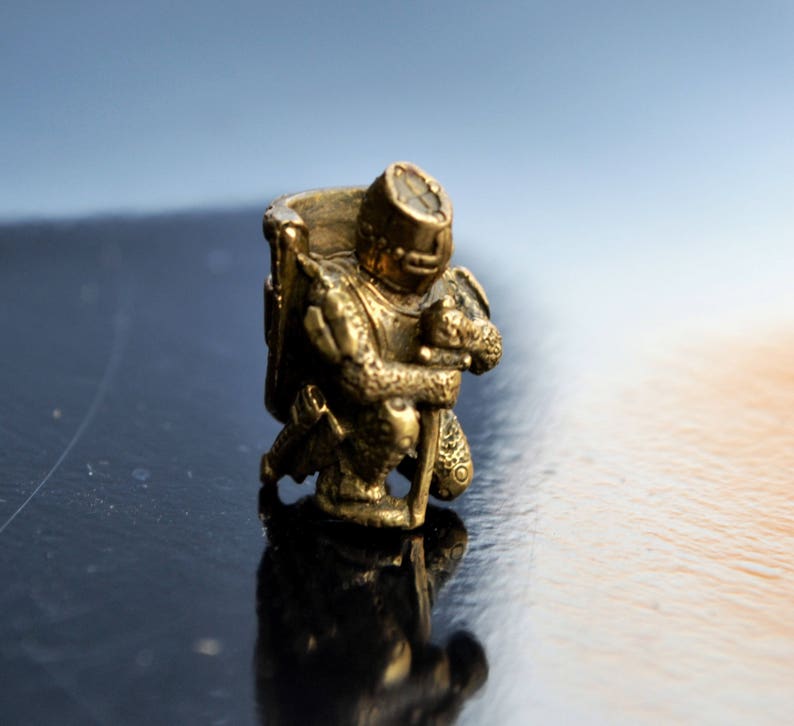 以下が含まれることがあります： 鎧を着たひざまずく騎士の小さな金色の置物。騎士は片手に剣、もう片手に盾を持っている。
