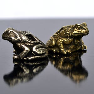 May include: Two small frog figurines, one gold and one silver, are sitting on a black surface. The frogs are facing forward with their mouths closed.