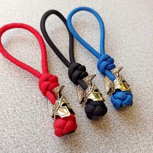 Op de afbeelding: Drie sleutelhangers met een gouden helmbedel. De sleutelhangers zijn gemaakt van rood, zwart en blauw koord.