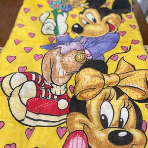 May include: Yellow sleeping bag featuring Mickey and Minnie Mouse. Mickey is wearing a purple jacket and holding flowers. Minnie has a yellow bow. The background is yellow with pink hearts. The sleeping bag is rectangular.