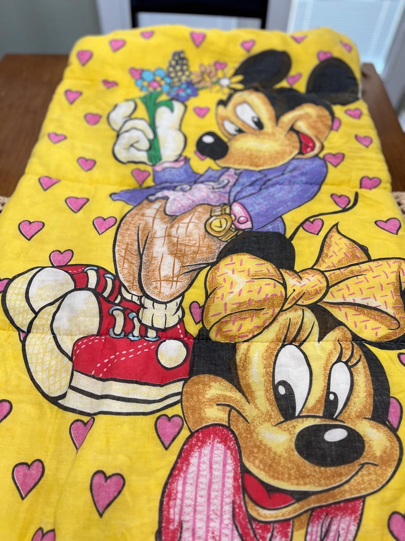 May include: Yellow sleeping bag featuring Mickey and Minnie Mouse. Mickey is wearing a purple jacket and holding flowers. Minnie has a yellow bow. The background is yellow with pink hearts. The sleeping bag is rectangular.
