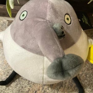 May include: A plush toy shaped like a bird with a long, gray beak and a round body. The bird has a gray head, white body, and black legs. The eyes are green with black pupils. A yellow tag is visible.