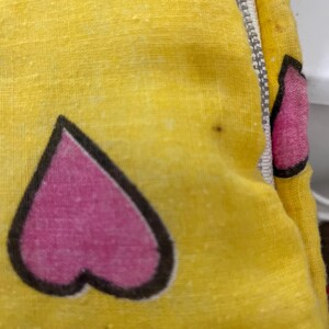 May include: Close-up of a yellow fabric with a repeating pattern of pink hearts outlined in black. A silver zipper runs along the edge of the fabric. The fabric has a slightly textured appearance.