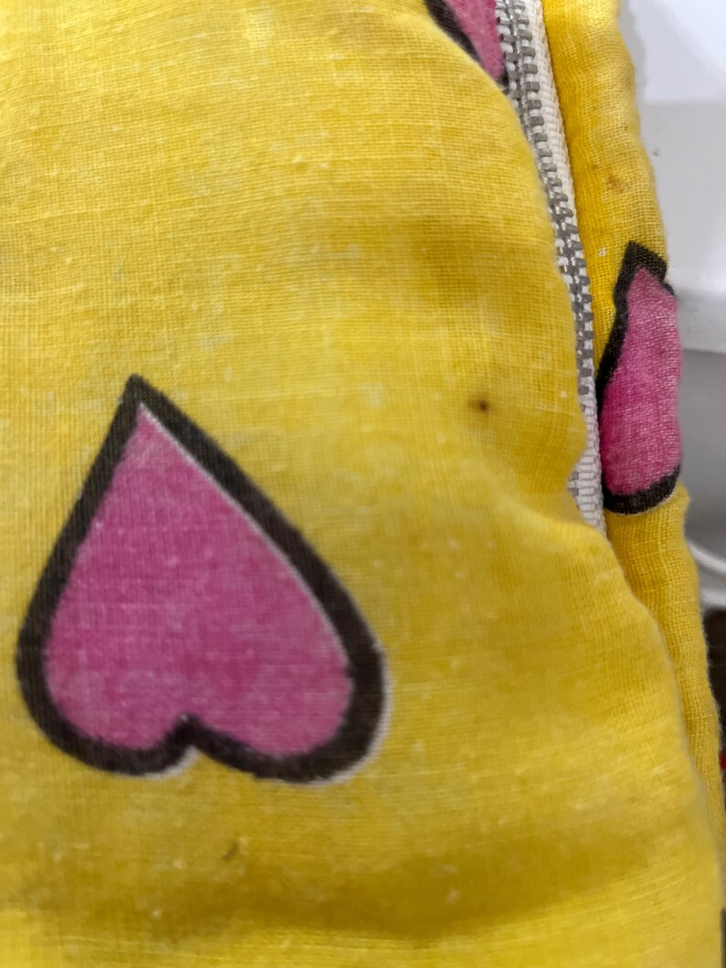 May include: Close-up of a yellow fabric with a repeating pattern of pink hearts outlined in black. A silver zipper runs along the edge of the fabric. The fabric has a slightly textured appearance.