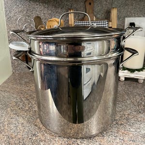 May include: A stainless steel Cuisinart stockpot with a matching lid and side handles. The pot features a polished, reflective finish and a cylindrical body. The brand name Cuisinart is clearly engraved on the front of the upper section of the pot.