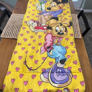 May include: Yellow sleeping bag featuring Mickey and Minnie Mouse characters. The bag has a repeating pattern of pink hearts. Mickey is wearing a purple jacket and red shoes. Minnie is wearing a pink outfit and holding a blue watering can.