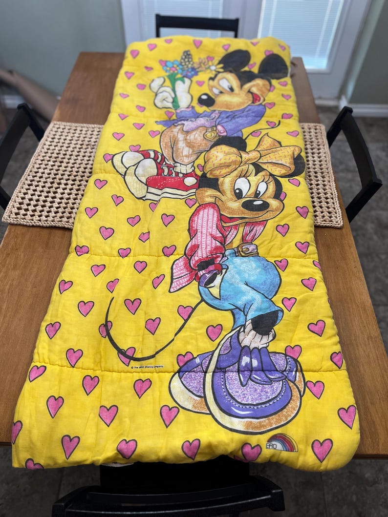 May include: Yellow sleeping bag featuring Mickey and Minnie Mouse characters. The bag has a repeating pattern of pink hearts. Mickey is wearing a purple jacket and red shoes. Minnie is wearing a pink outfit and holding a blue watering can.