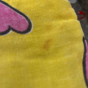 May include: Close-up of a yellow fabric with a pink heart-shaped design outlined in black. The fabric has a few brown stains and small white specks. The image is of a pillow or cushion.