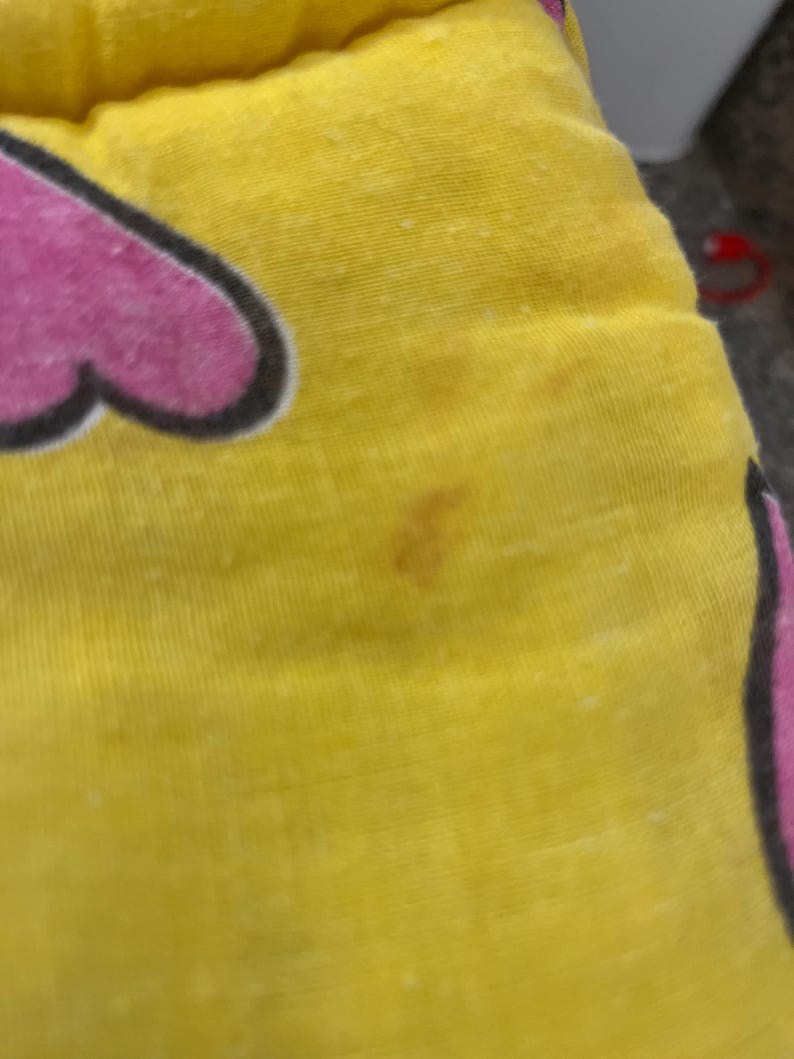May include: Close-up of a yellow fabric with a pink heart-shaped design outlined in black. The fabric has a few brown stains and small white specks. The image is of a pillow or cushion.