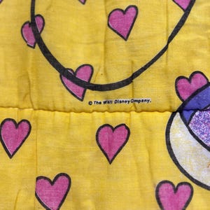 May include: Close-up of a yellow fabric with a pattern of pink hearts outlined in black. A black curved line is present, and the text "The Walt Disney Company" is visible.