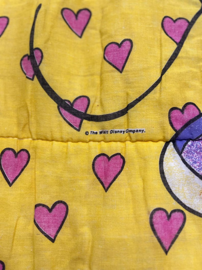 May include: Close-up of a yellow fabric with a pattern of pink hearts outlined in black. A black curved line is present, and the text "The Walt Disney Company" is visible.